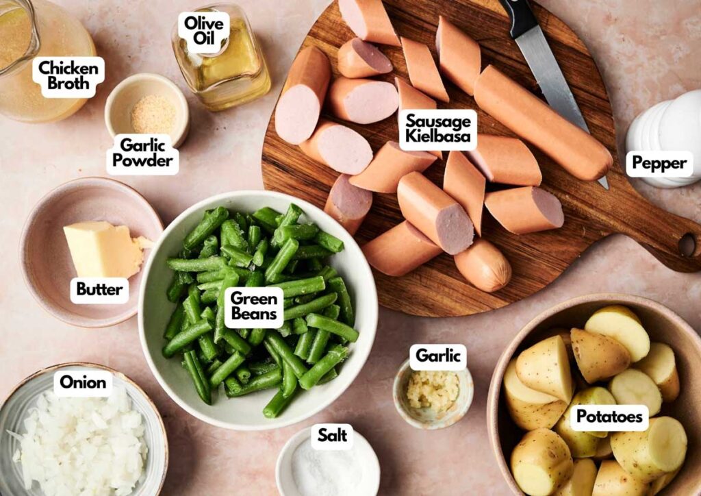 Ingredients for a Sausage Green Bean Potato Casserole, including sliced kielbasa, potatoes, green beans, butter, garlic, onion, chicken broth, olive oil, and seasonings are displayed on the countertop.