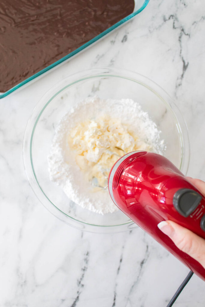 A person uses a red hand mixer to blend powdered sugar and a creamy ingredient in a glass bowl, preparing the topping for Strawberry Cheesecake Brownies; a chocolate dessert sits in a baking dish nearby.