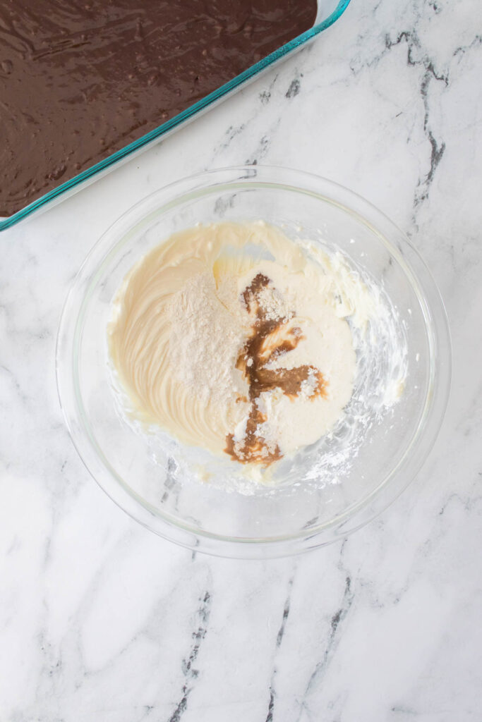 A glass bowl with cream, flour, and vanilla mixture sits on a marble countertop next to a baking dish with chocolate batter, ready to create delicious Strawberry Cheesecake Brownies.