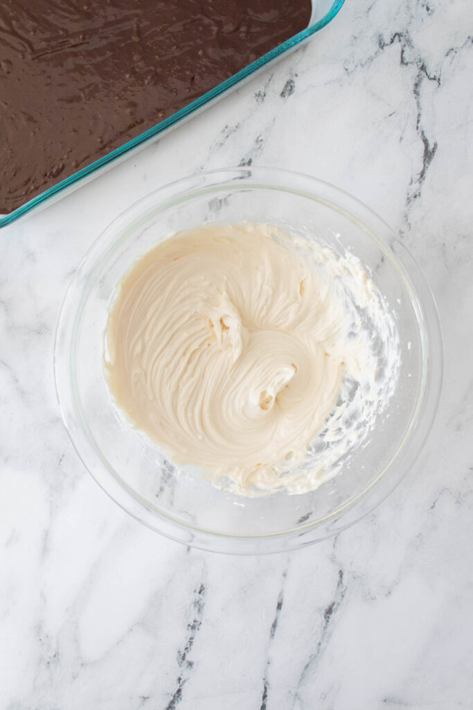 A glass bowl with creamy white frosting sits on a marble countertop; a glass dish with Strawberry Cheesecake Brownies is partially visible in the upper left corner.