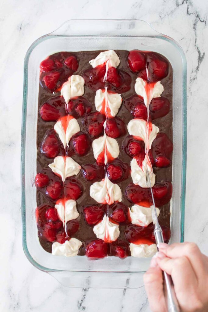 A hand uses a knife to swirl cream cheese and cherry topping into brownie batter in a glass baking dish on a marble surface, creating a Strawberry Cheesecake Brownies-inspired treat.