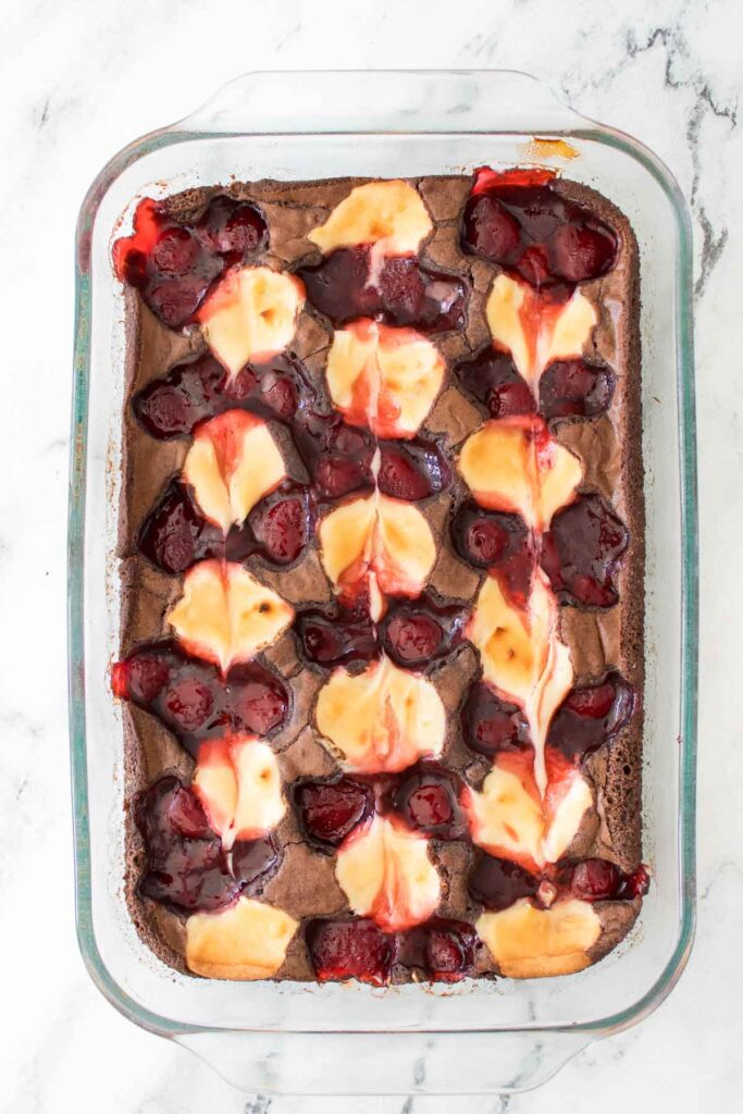 A glass baking dish filled with Strawberry Cheesecake Brownies, topped with swirls of creamy cheesecake and cherry pie filling, rests on a white marble surface.