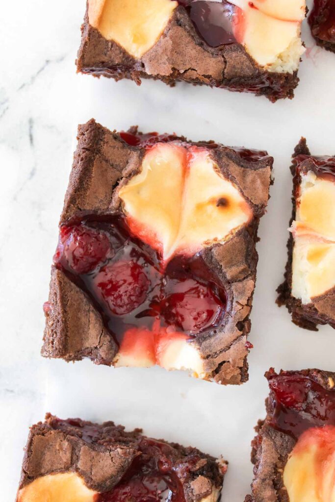 A close-up of Strawberry Cheesecake Brownies, cut into squares and topped with swirls of cheesecake and cherry filling, arranged on a white surface.