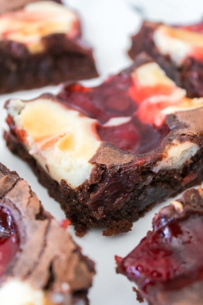 A close-up of Strawberry Cheesecake Brownies with a swirl of cherry filling on top, cut into squares and arranged on a white surface.