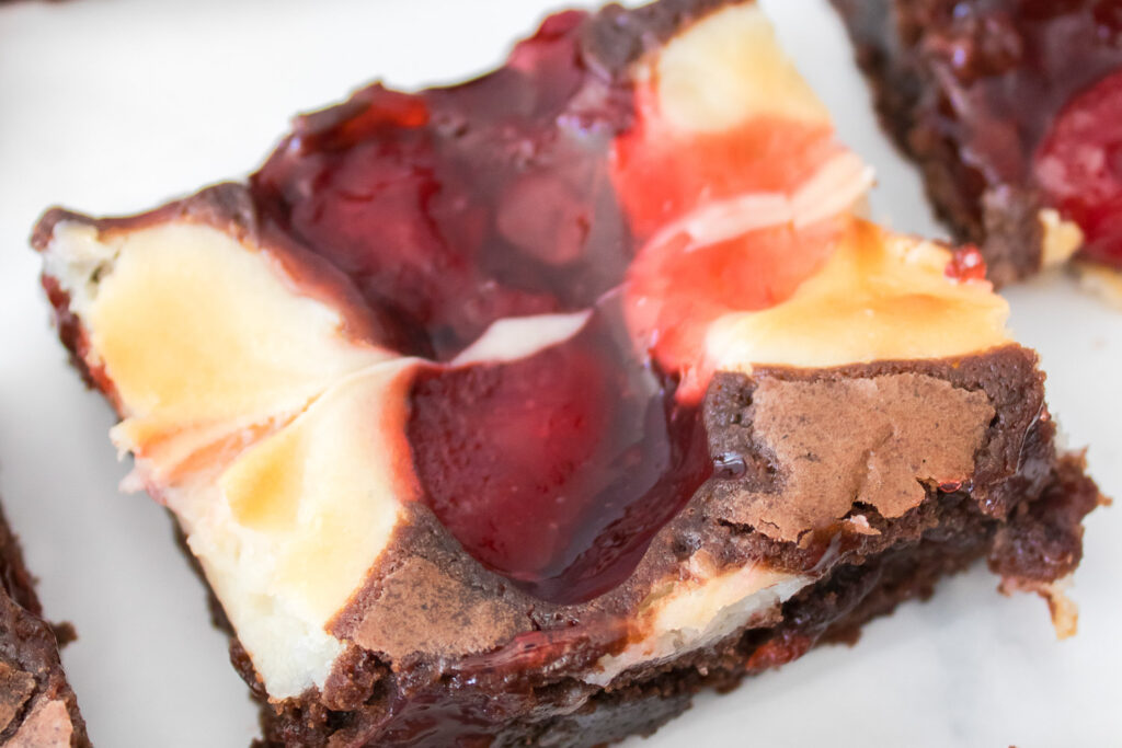 A close-up of Strawberry Cheesecake Brownies, topped with swirls of cream cheese and strawberry jam, on a white surface.