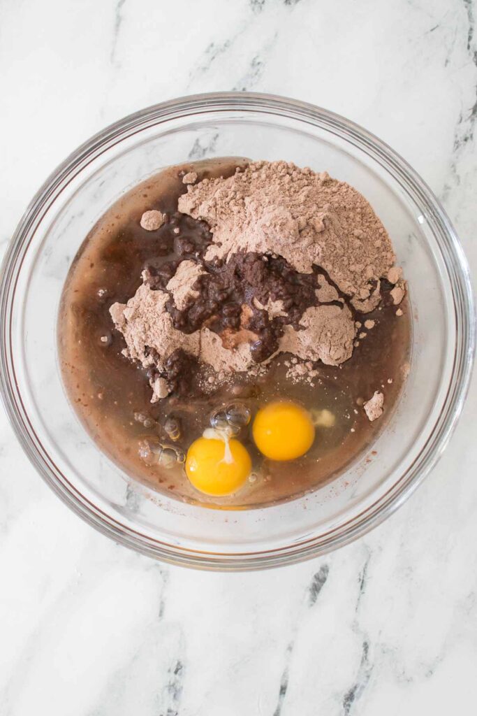 A glass bowl containing Strawberry Cheesecake Brownies mix powder, two raw eggs, and water on a white marble surface.