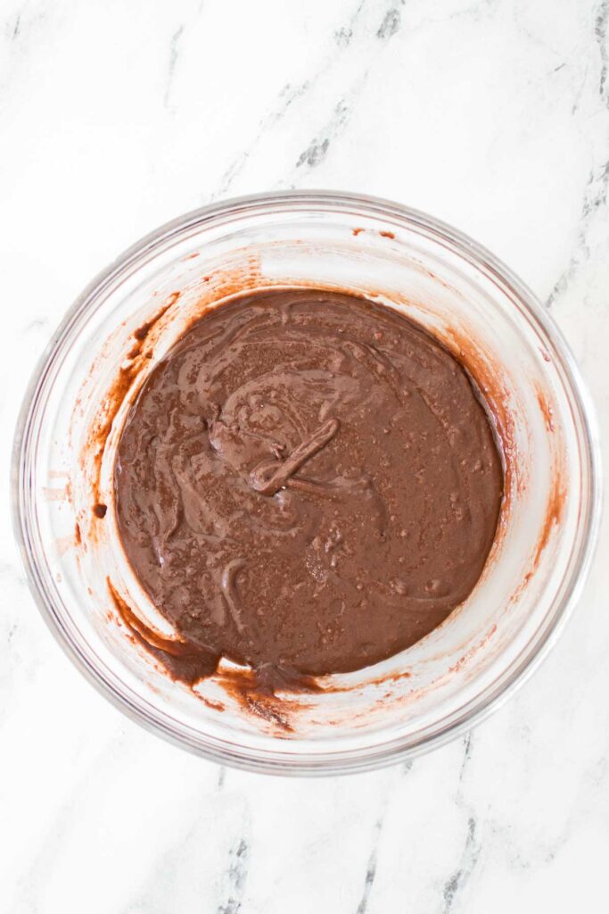 A clear glass bowl filled with rich Strawberry Cheesecake Brownie batter sits on a white marble surface.