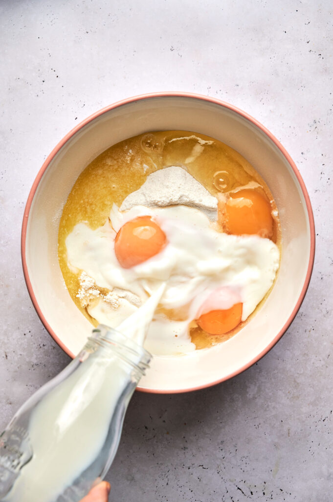 A bowl contains flour, eggs, and melted butter, with milk being poured in from a glass bottle—perfect beginnings for a delicious Strawberry Poke Cake.