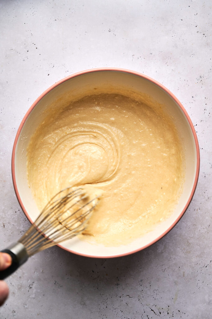A hand is whisking a bowl of pale yellow batter on a light-colored surface, preparing the base for a delicious Strawberry Poke Cake.
