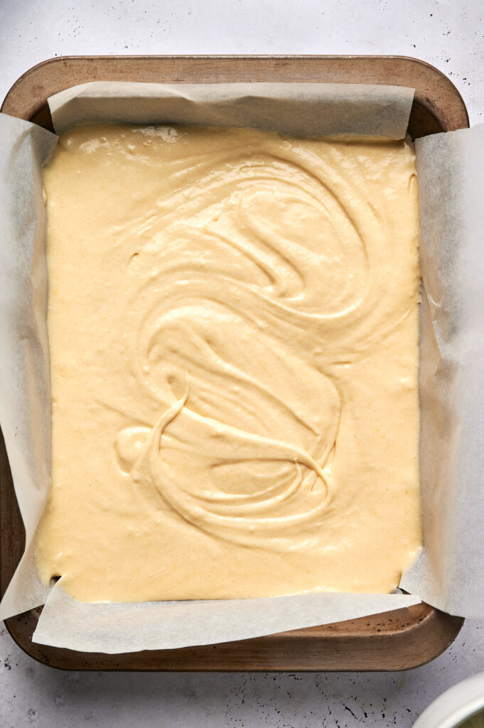 A baking tray lined with parchment paper is filled with smooth, pale yellow Strawberry Poke Cake batter, ready to be baked.