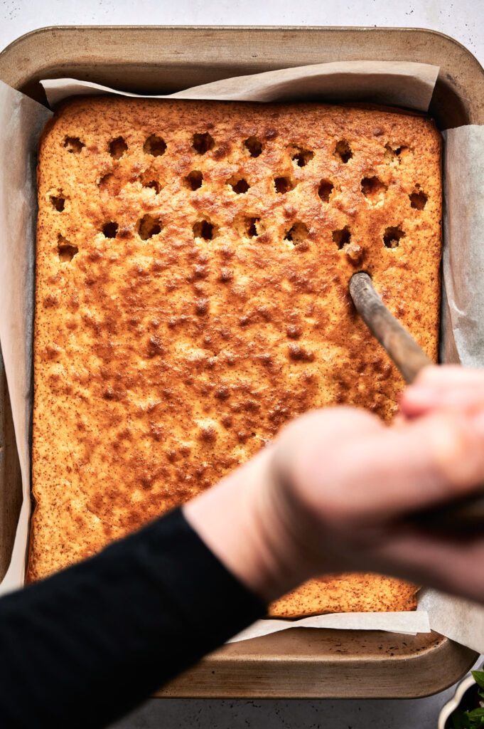 A hand uses a wooden spoon to poke holes into a rectangular Strawberry Poke Cake in a parchment-lined pan.