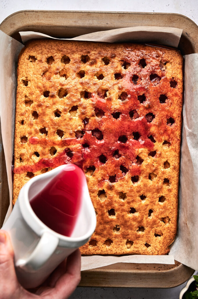 A hand pours red liquid over a rectangular Strawberry Poke Cake with evenly spaced holes, set in a parchment-lined baking pan.