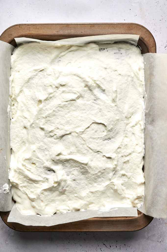 A parchment-lined baking pan filled with a layer of whipped cream spread evenly over a delicious Strawberry Poke Cake dessert.