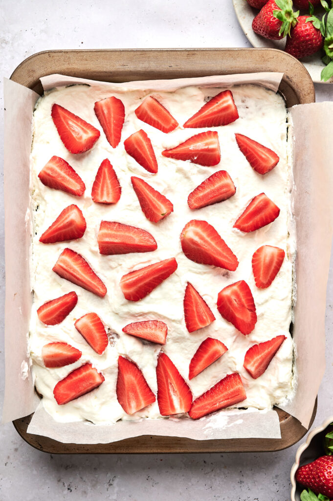 A square baking pan lined with parchment paper, filled with whipped cream and topped with sliced fresh strawberries, perfect for making a delicious Strawberry Poke Cake.