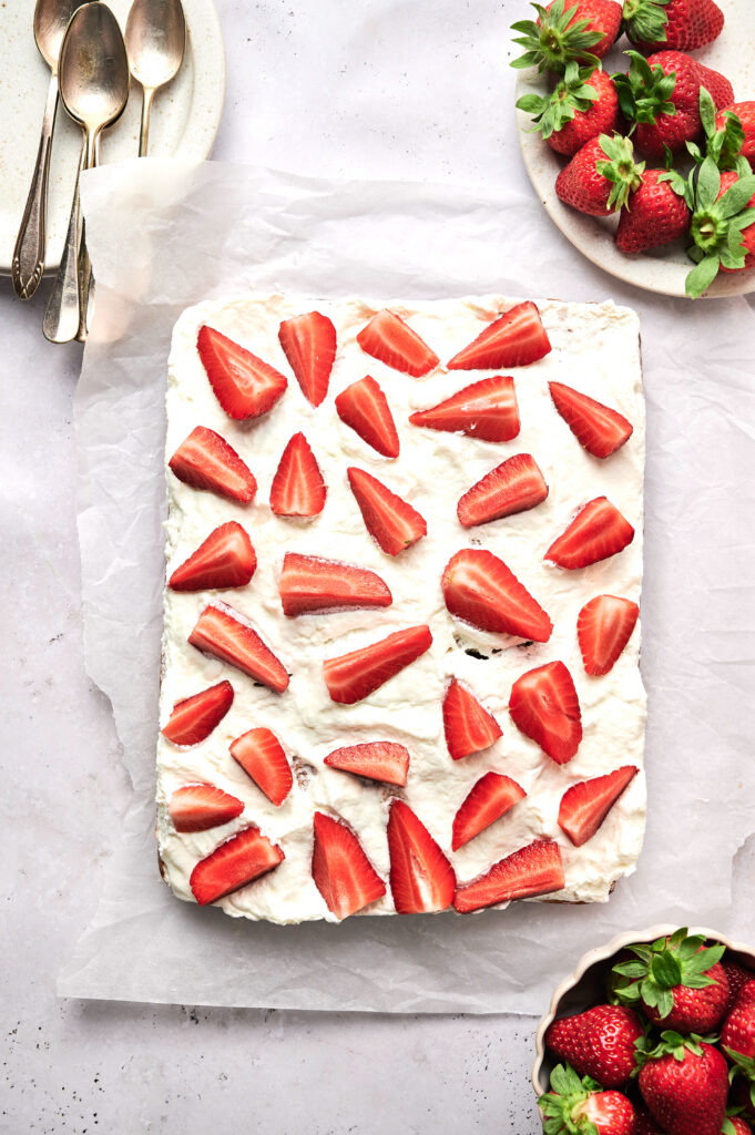 Rectangular Strawberry Poke Cake with white frosting, topped with halved strawberries, on parchment paper. Spoons and bowls of fresh strawberries are placed nearby.