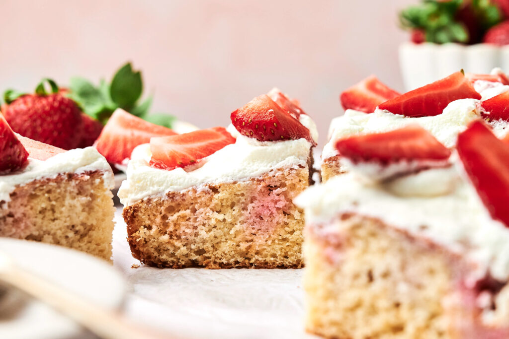 Slices of Strawberry Poke Cake topped with whipped cream and fresh strawberry pieces, with whole strawberries in the background.