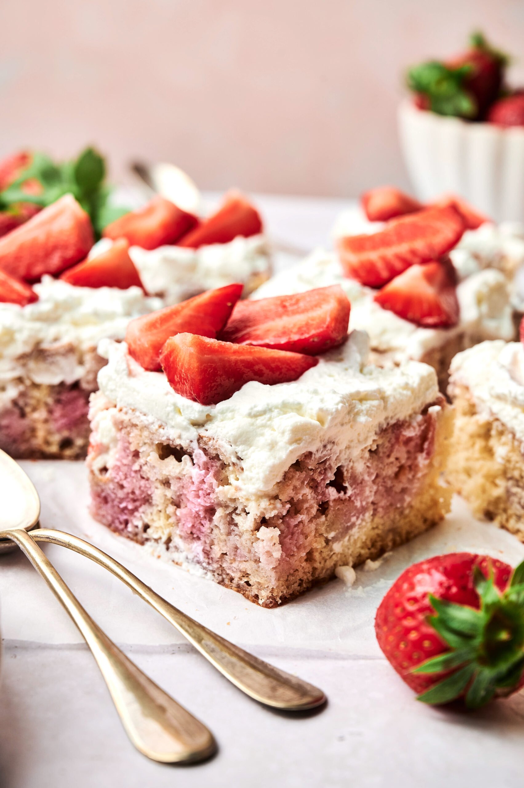 Three square pieces of Strawberry Poke Cake with white frosting and fresh strawberry slices on top, placed on a white surface with spoons and whole strawberries nearby.