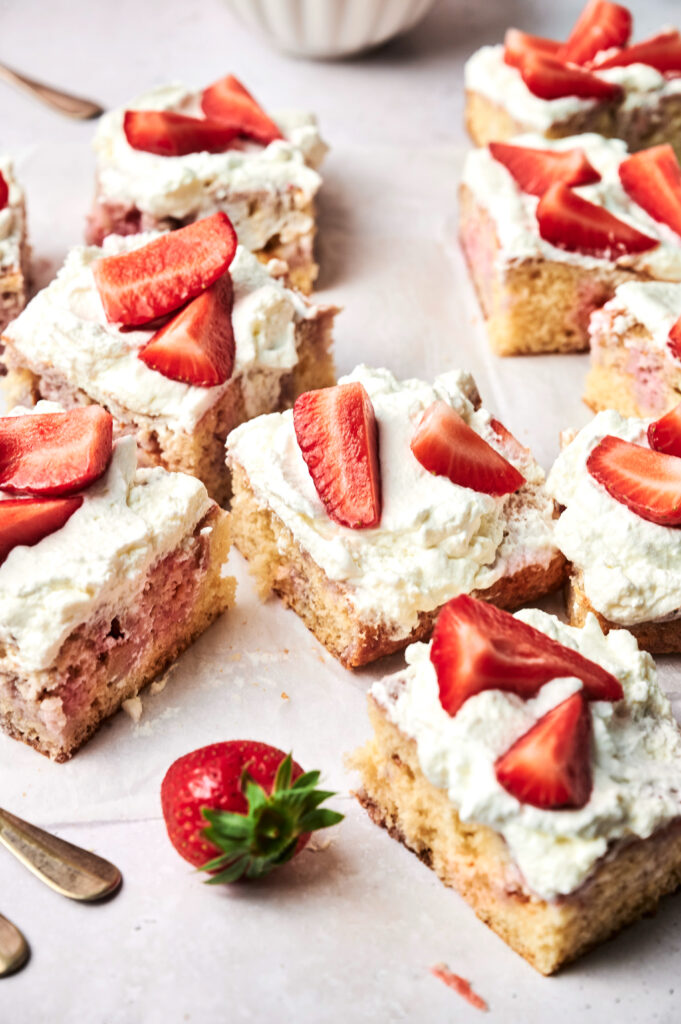Squares of Strawberry Poke Cake topped with whipped cream and sliced strawberries are arranged on a light surface, with a whole strawberry nearby.