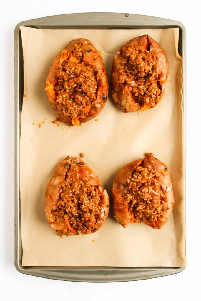 Four baked sweet potatoes stuffed with a savory Sweet Potato Sloppy Joes ground meat mixture are arranged on a parchment-lined baking sheet.