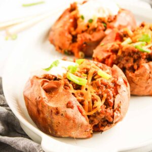 Baked sweet potatoes split open and filled with Sweet Potato Sloppy Joes mixture, shredded cheese, and a dollop of sour cream, served on a white plate.
