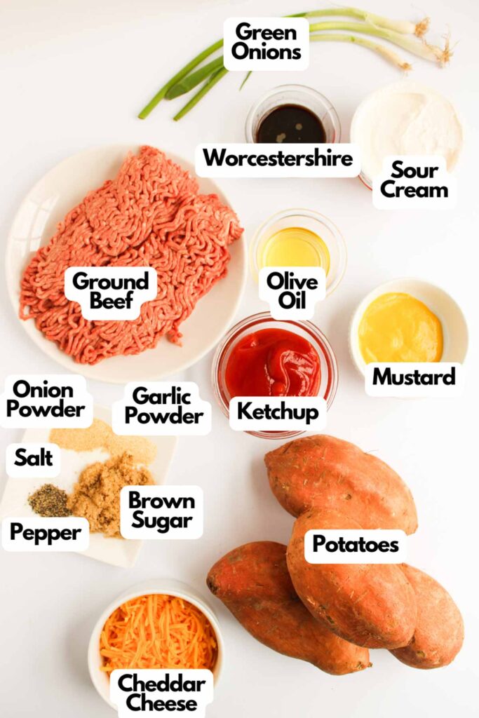 Various labeled ingredients for Sweet Potato Sloppy Joes are arranged on a white surface, including ground beef, sweet potatoes, seasonings, sauces, sour cream, green onions, and cheddar cheese.