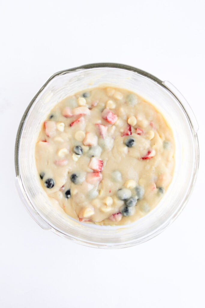 A glass mixing bowl filled with 4th of July Muffins batter—blueberries, chopped strawberries, and white chocolate chips—viewed from above on a white background.