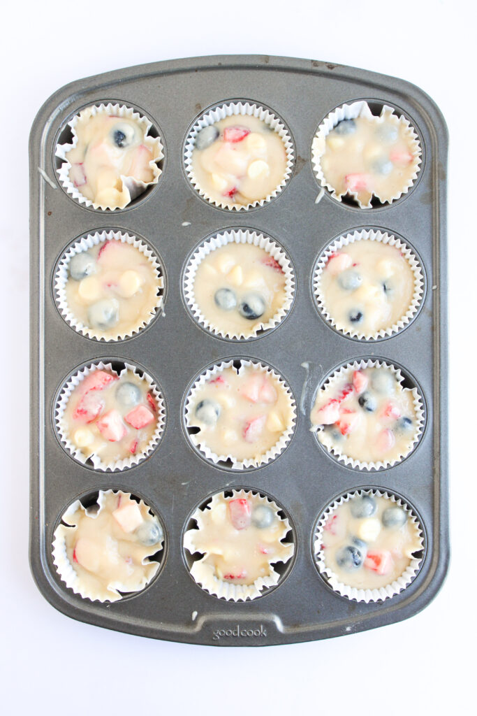 A muffin tin with twelve paper liners filled with uncooked batter containing visible blueberries and chopped strawberries, perfect for making festive 4th of July Muffins.