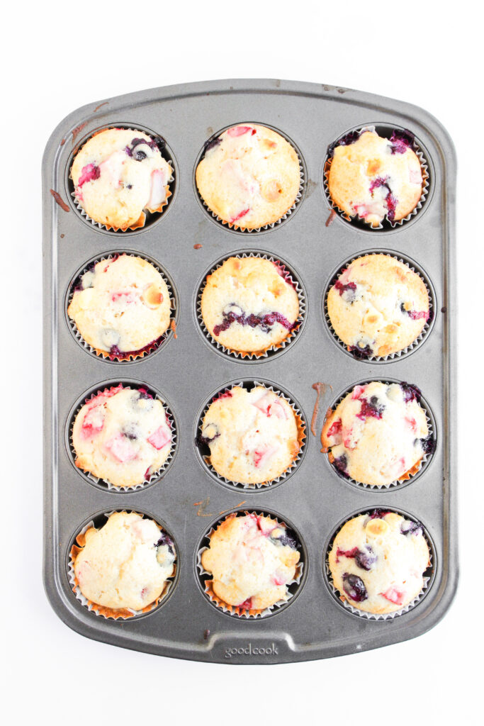 A muffin tin containing twelve baked 4th of July Muffins with visible blueberries and strawberries on top, viewed from above against a white background.