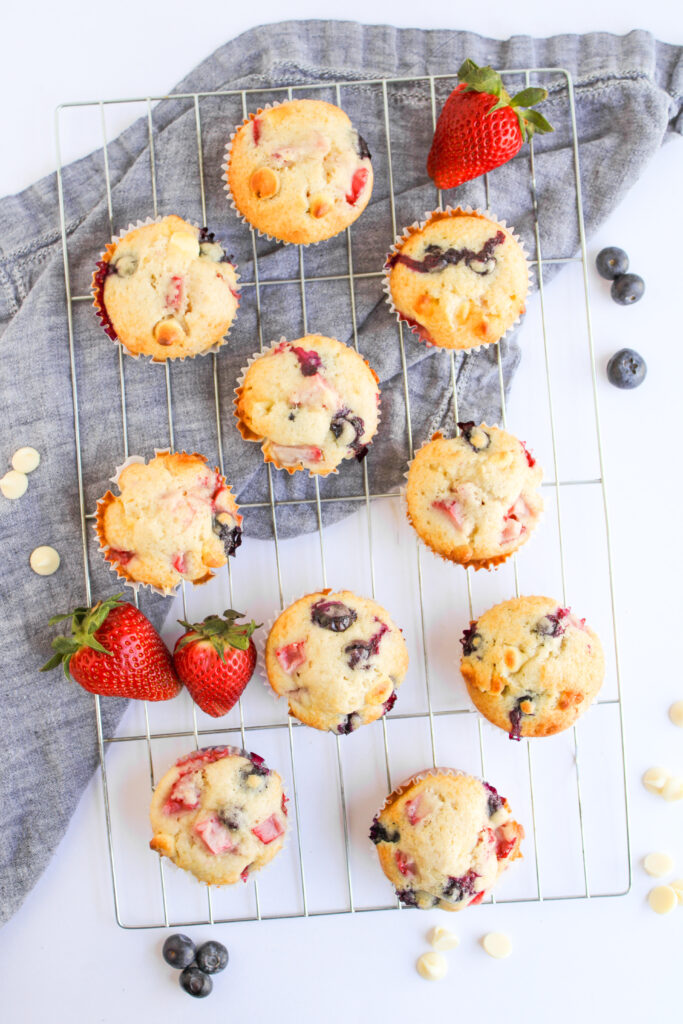 A cooling rack with assorted 4th of July Muffins, fresh strawberries, blueberries, and white chocolate chips on a grey cloth.