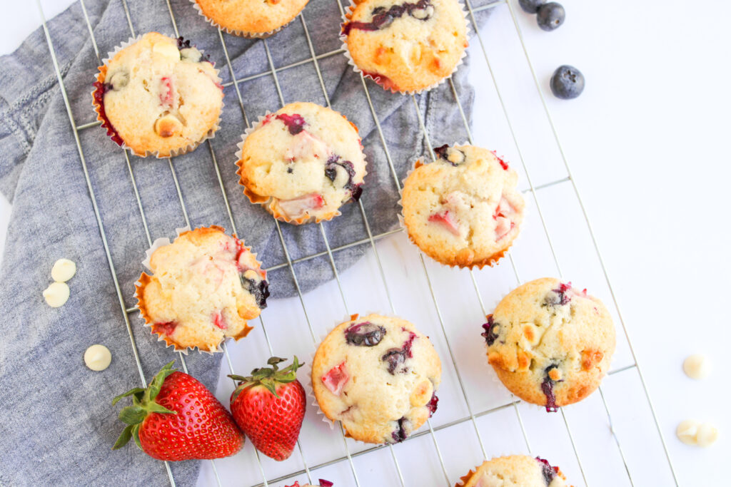 4th of July Muffins with blueberries and strawberries cool on a wire rack, surrounded by fresh strawberries, blueberries, and white chocolate chips.