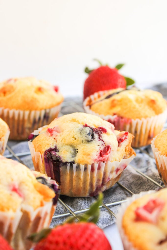 Assorted 4th of July Muffins in paper liners on a cooling rack, with whole strawberries placed nearby for a festive touch.