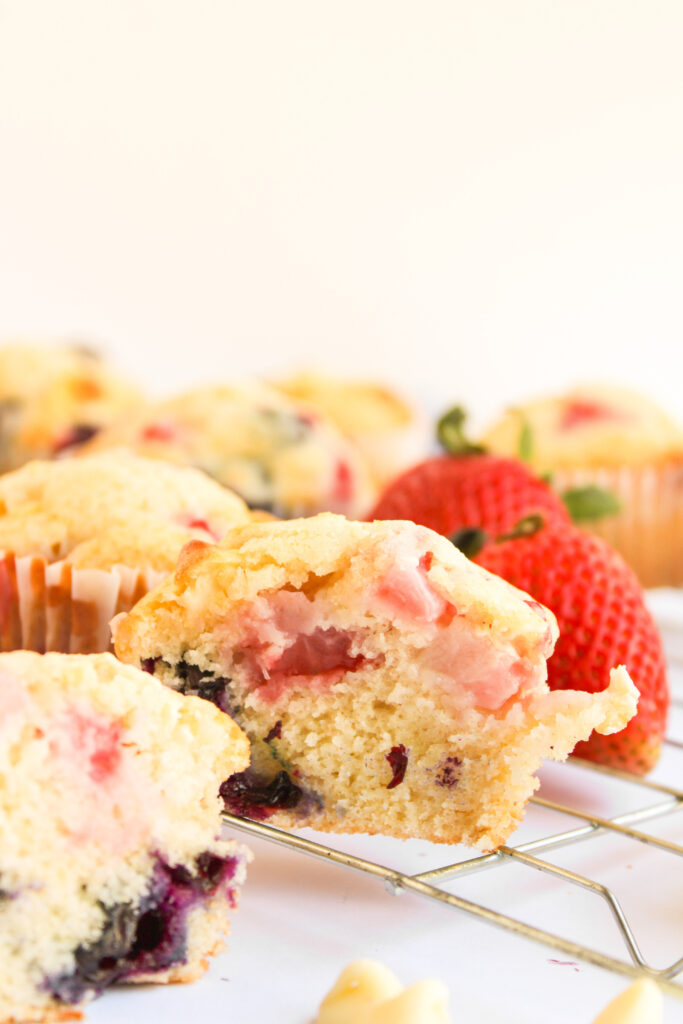A partially eaten berry muffin sits on a cooling rack next to a whole strawberry, with more 4th of July Muffins visible in the background.