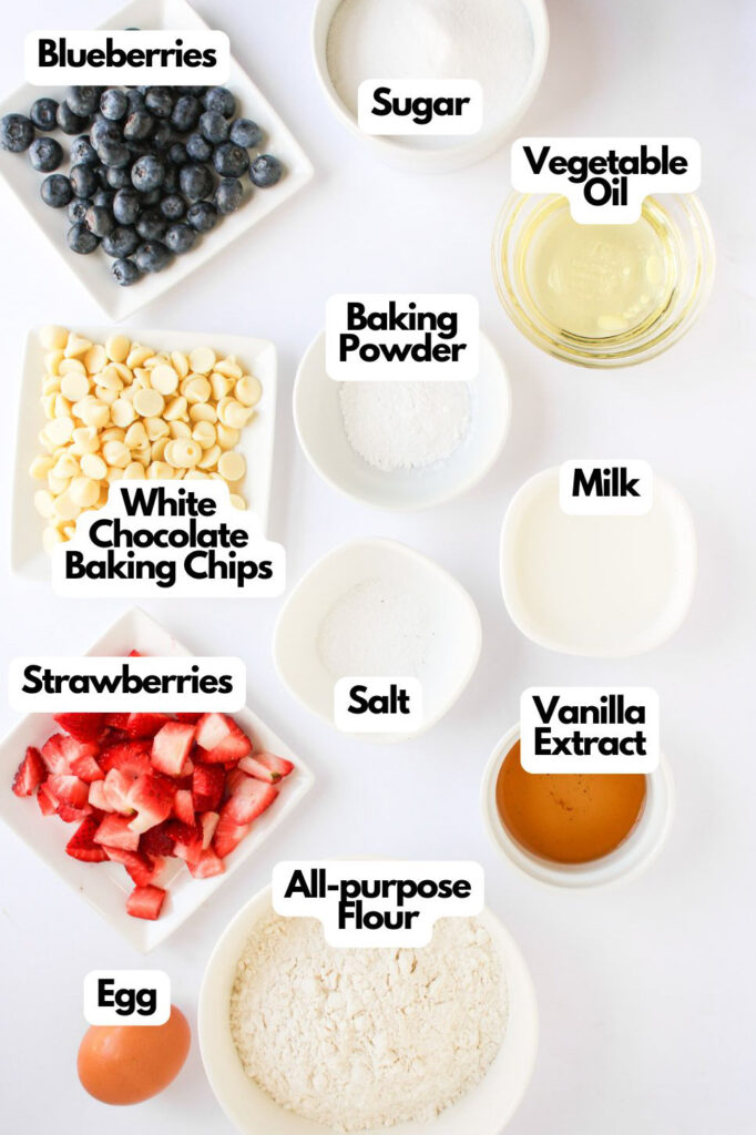 Flat lay of baking ingredients for 4th of July Muffins in labeled bowls: blueberries, strawberries, white chocolate chips, egg, sugar, oil, milk, flour, baking powder, salt, and vanilla extract.