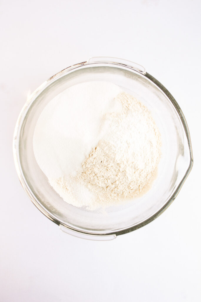 A glass mixing bowl containing separate piles of flour and granulated sugar on a white background, perfect for preparing festive 4th of July Muffins.
