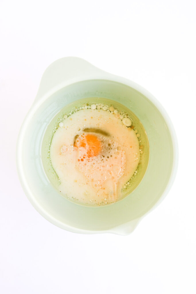 A cracked egg, milk, and other liquid ingredients for 4th of July Muffins sit in a light green mixing bowl, viewed from above, against a white background.