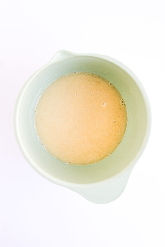 A light green mixing bowl filled with a pale, frothy liquid mixture for 4th of July Muffins, viewed from above on a white background.