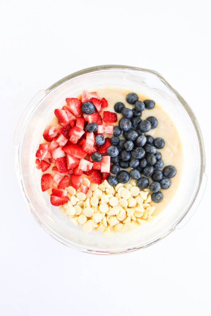 A glass bowl contains muffin batter topped with chopped strawberries, blueberries, and white chocolate chips, viewed from above—perfect for festive 4th of July Muffins.