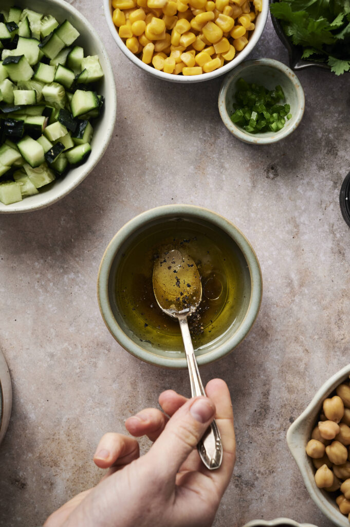 A hand stirs olive oil and pepper in a bowl, surrounded by bowls of chopped cucumber, corn, green chili, chickpeas, and cilantro&mdash;perfect fresh ingredients for making cowboy caviar on the countertop.