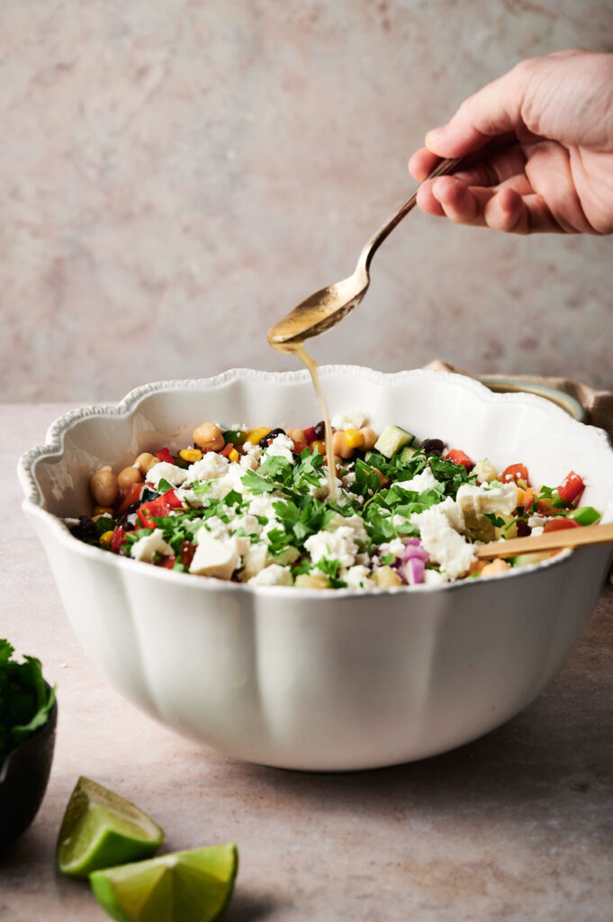 A hand drizzles dressing from a spoon over a cowboy caviar-inspired salad with feta, chickpeas, cucumber, tomato, and herbs in a white bowl; lime wedges sit nearby.