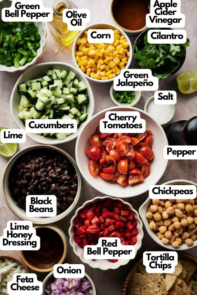 A variety of labeled ingredients for a salad, including chopped vegetables, beans, cheese, chips, dressing, and seasonings&mdash;perfect for assembling your own cowboy caviar&mdash;are arranged in bowls on a table.