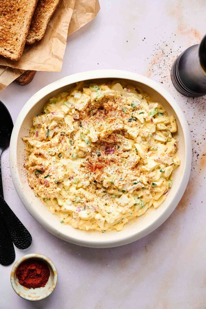 A bowl of creamy egg salad topped with paprika and herbs, served with slices of toast and a small bowl of paprika on a light surface.