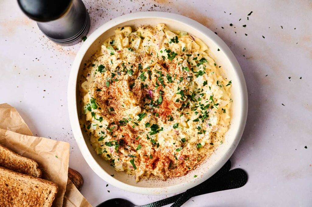 A bowl of creamy egg salad topped with chopped herbs and paprika, served alongside slices of toast and a pepper grinder on a light surface.