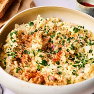 A bowl of potato and egg salad topped with chopped parsley and paprika, with slices of toasted bread in the background.