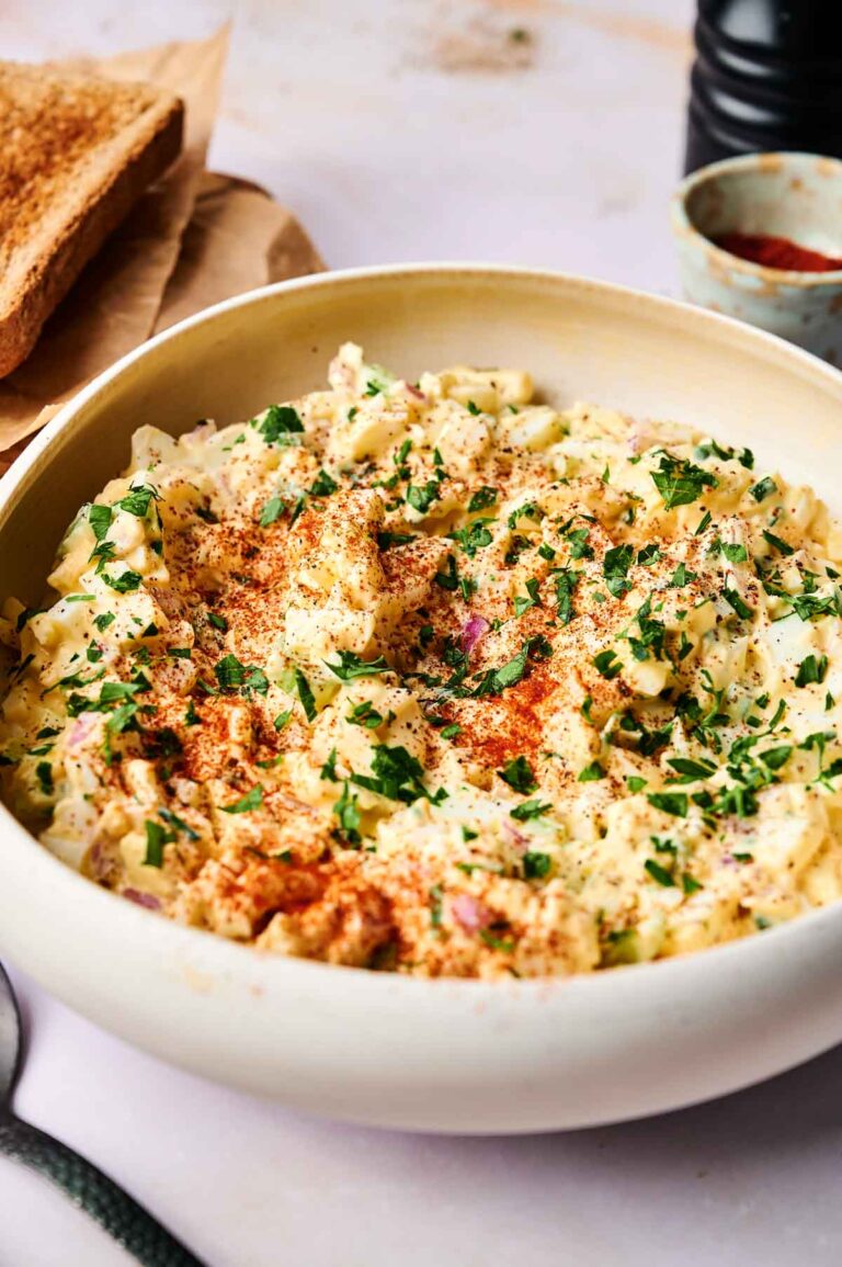 A bowl of potato and egg salad topped with chopped parsley and paprika, with slices of toasted bread in the background.