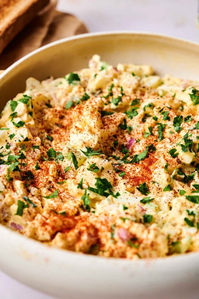 A bowl of potato and egg salad topped with chopped parsley and paprika.