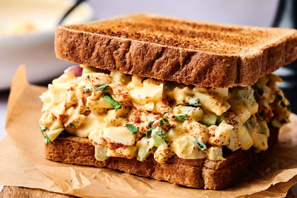 A close-up of an egg salad sandwich with chopped vegetables and herbs on toasted whole wheat bread, sprinkled with paprika, resting on brown parchment paper. This classic egg salad creation is both fresh and flavorful.