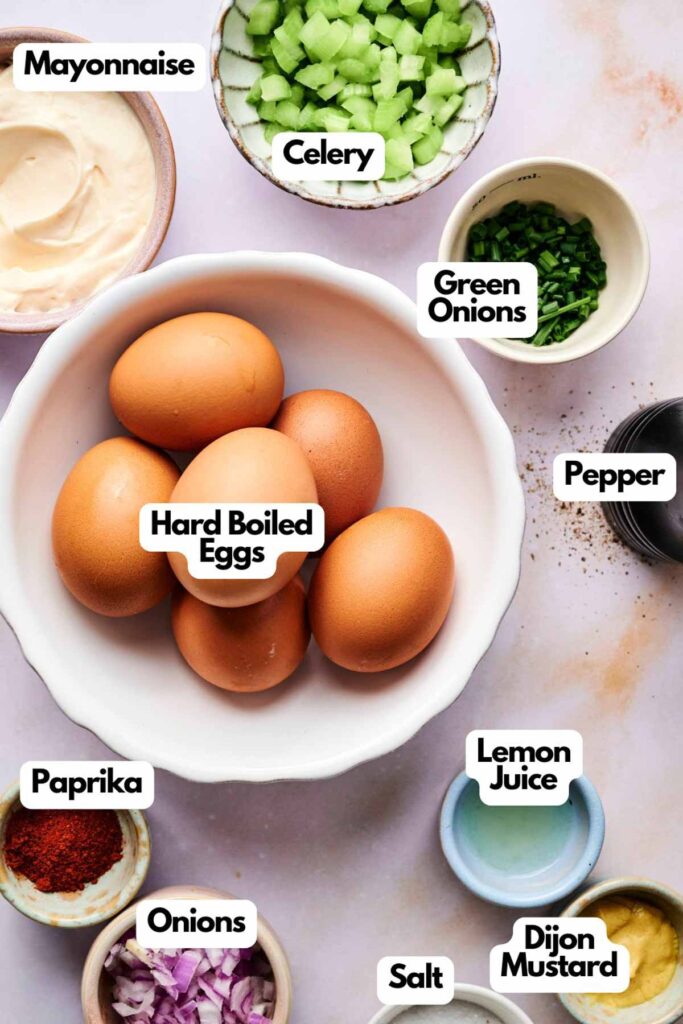 A bowl of hard boiled eggs sits ready for making Egg Salad, surrounded by labeled bowls of mayonnaise, celery, green onions, pepper, paprika, lemon juice, onions, salt, and Dijon mustard.