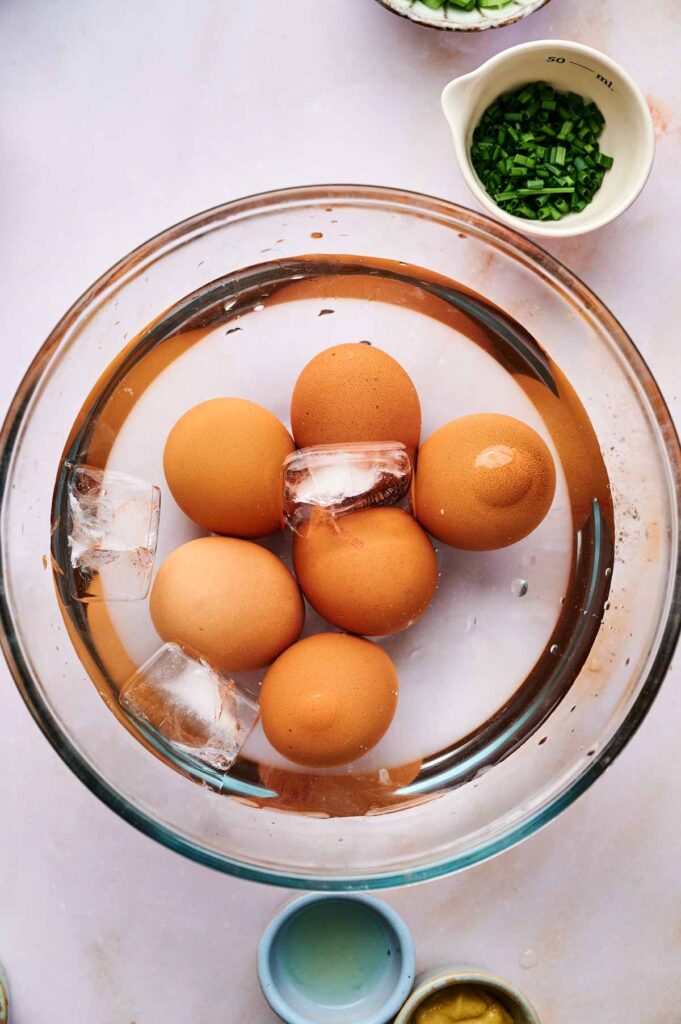 A glass bowl with six brown eggs and ice cubes in water, perfect for prepping Egg Salad, is surrounded by small bowls containing chopped herbs and condiments.