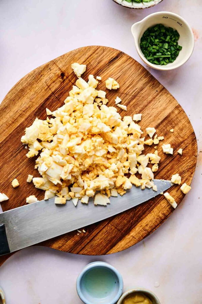 Chopped hard-boiled eggs on a wooden cutting board with a knife, surrounded by small bowls of herbs and condiments—perfect ingredients for making a fresh Egg Salad.