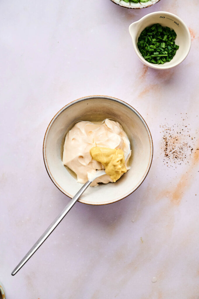 A bowl containing mayonnaise and mustard with a spoon, next to a small cup of chopped herbs on a light-colored surface—perfect ingredients for making a classic egg salad.